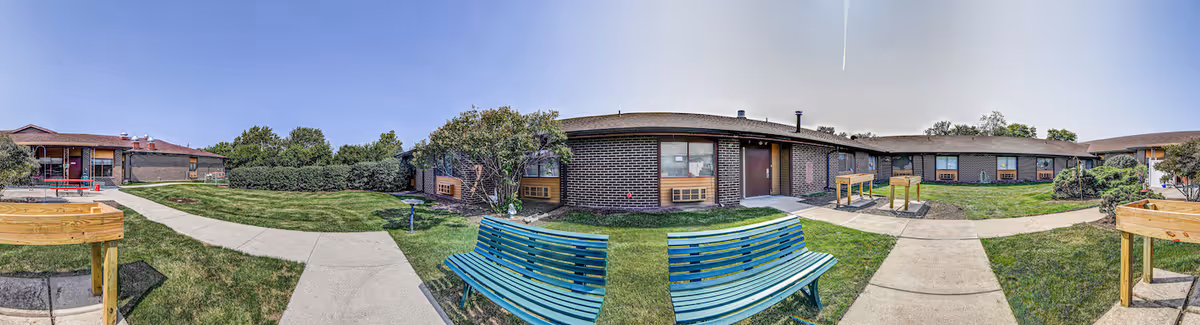 Panoramic outdoor view of a senior living facility courtyard with green grass, paved walkways, blue benches, and a brick building under a clear sky.