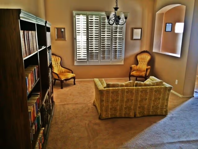 A cozy living room with a patterned sofa facing two yellow upholstered armchairs. There is a large window with white plantation shutters on the far wall, flanked by two framed pictures. A dark wooden bookshelf filled with books is on the left side. The room has beige walls and carpet, and a black chandelier with white lampshades hangs from the ceiling.