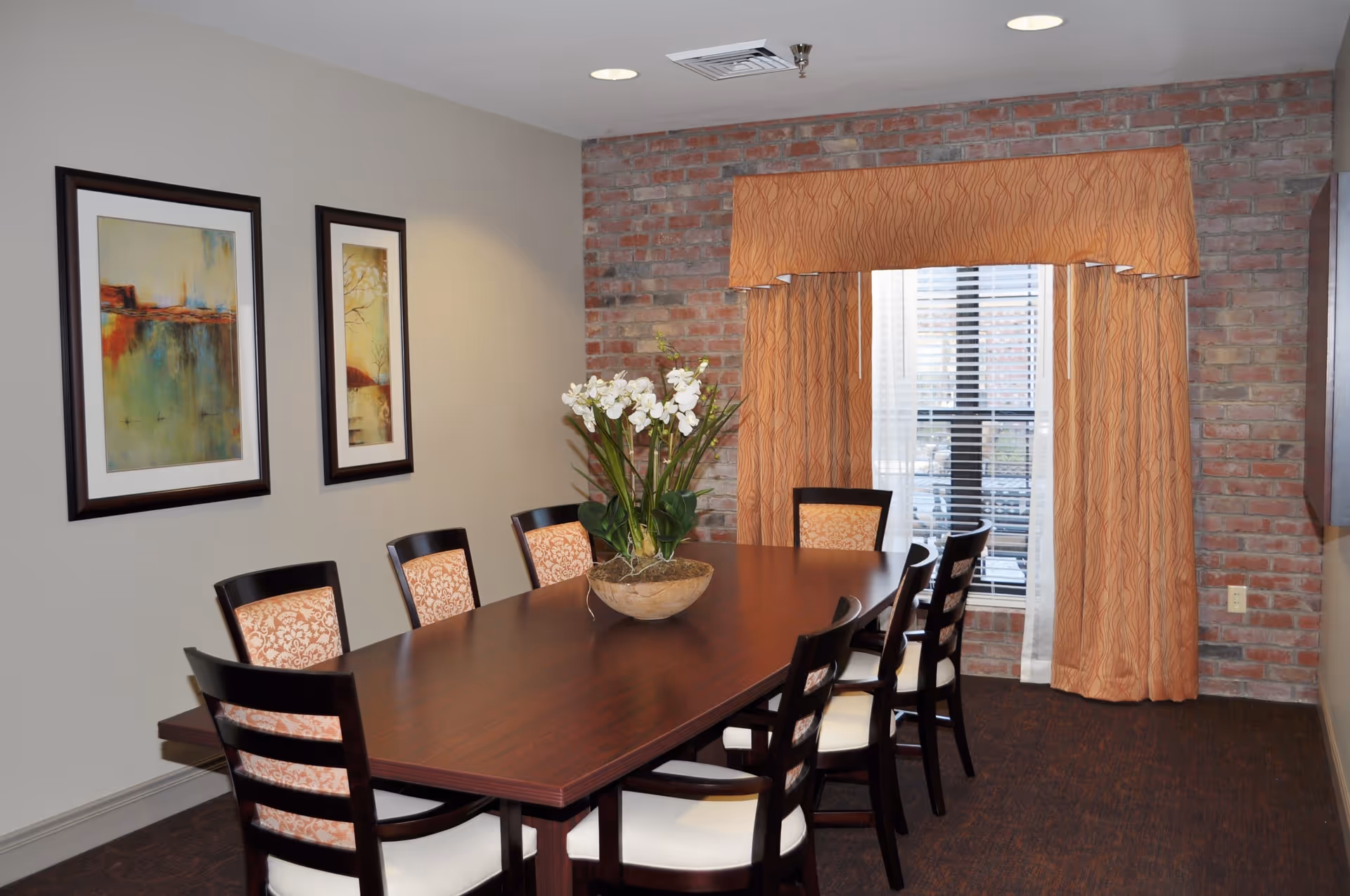 A formal dining room with a long wooden table surrounded by chairs, a floral centerpiece, framed artwork on the wall, and a window with orange drapes set against an exposed brick wall.