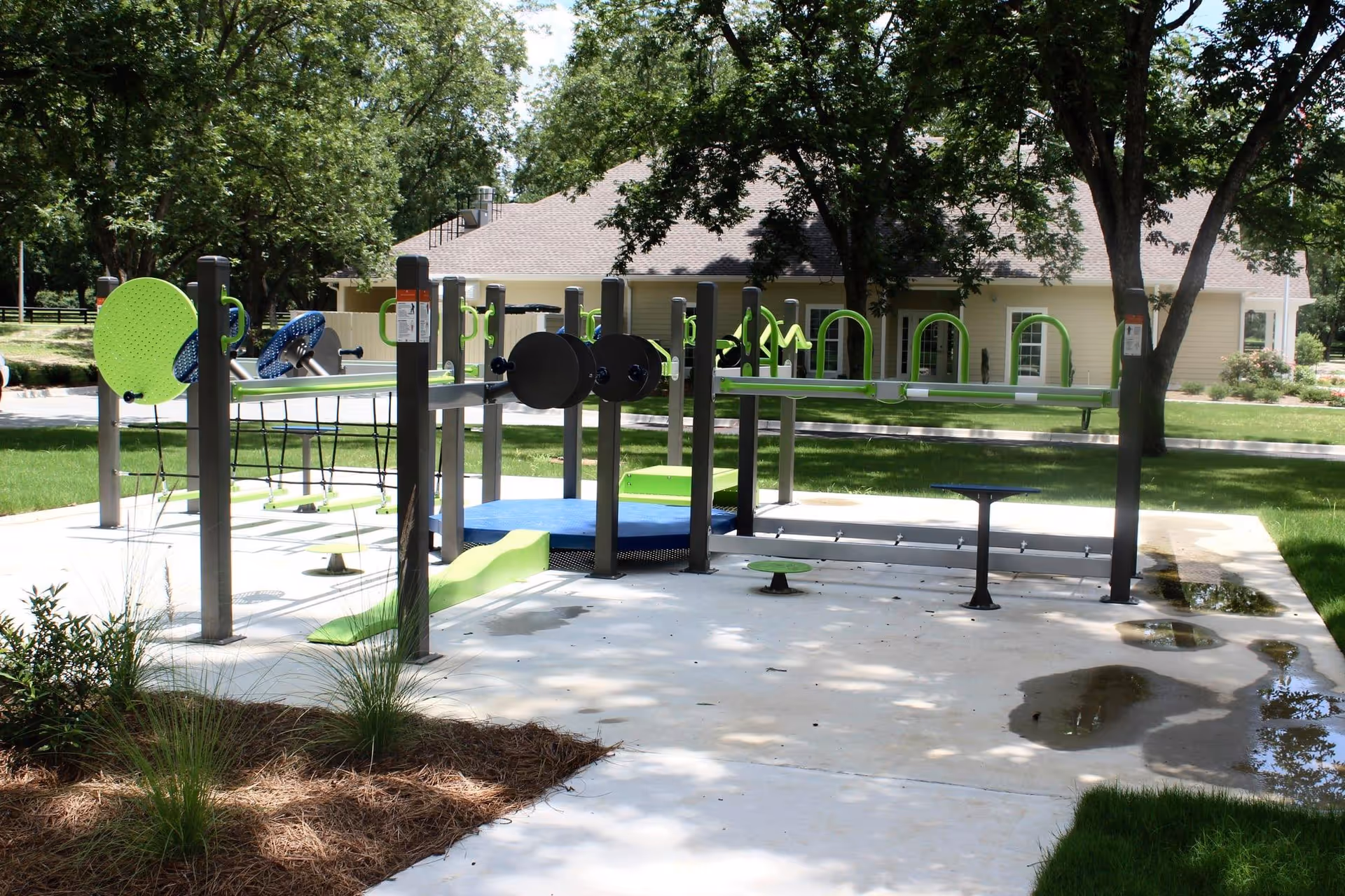 Outdoor exercise equipment in a park-like setting with trees and a building in the background under a sunny sky.