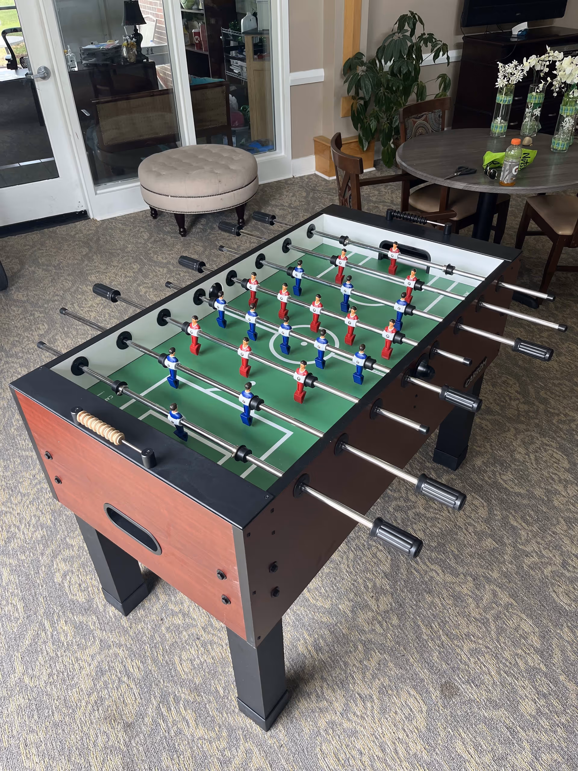 Foosball table with red and blue players set up in a room with carpeted floor. In the background, there is a round table with chairs, some decorative flowers in vases, a plant, and a beige round ottoman near glass doors.