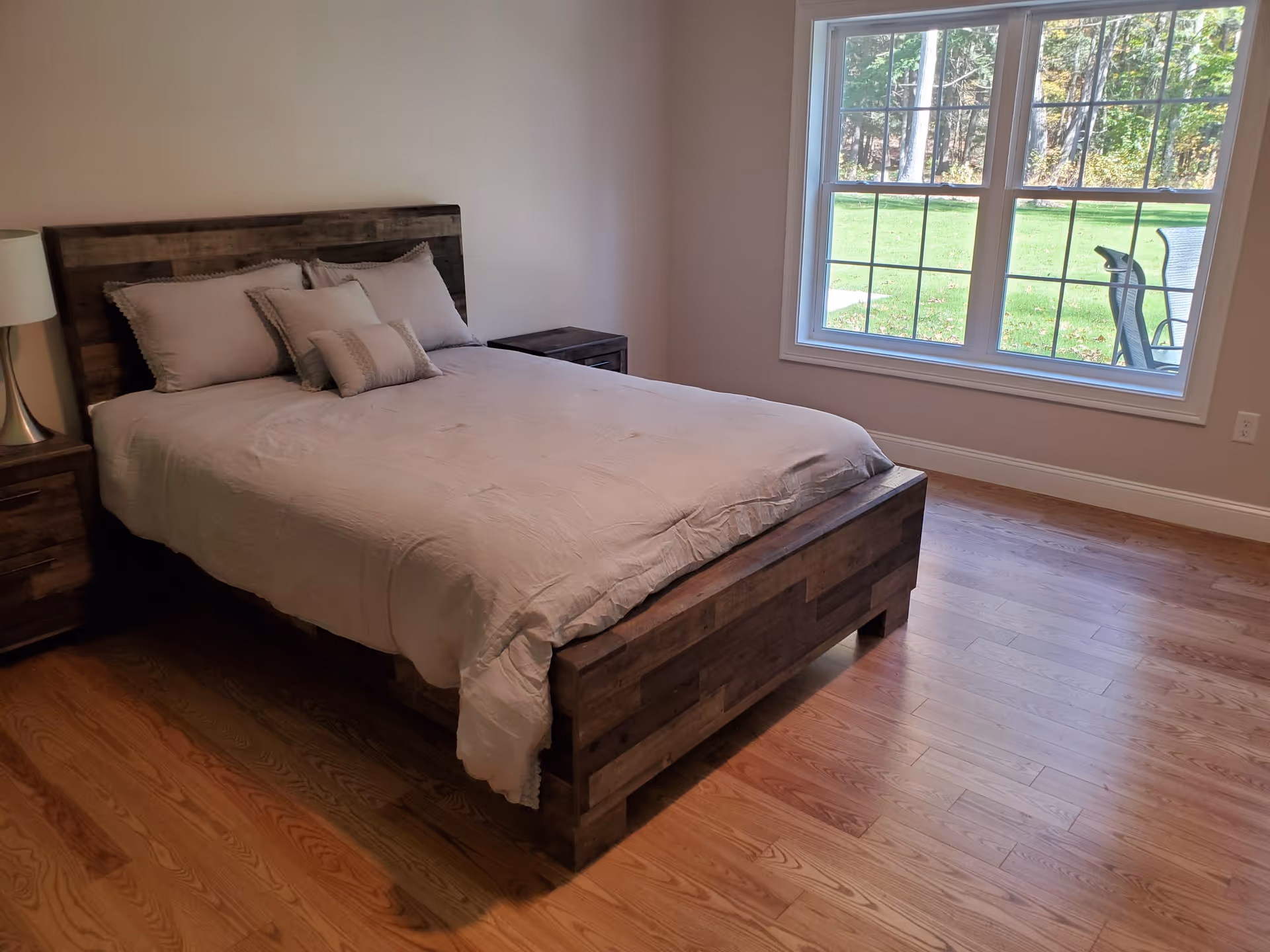 A bedroom with a wooden bed frame and headboard, made up with light-colored bedding and several pillows. There are two matching wooden nightstands on either side of the bed, one with a lamp. A large window shows a view of green grass and trees outside. The floor is hardwood.