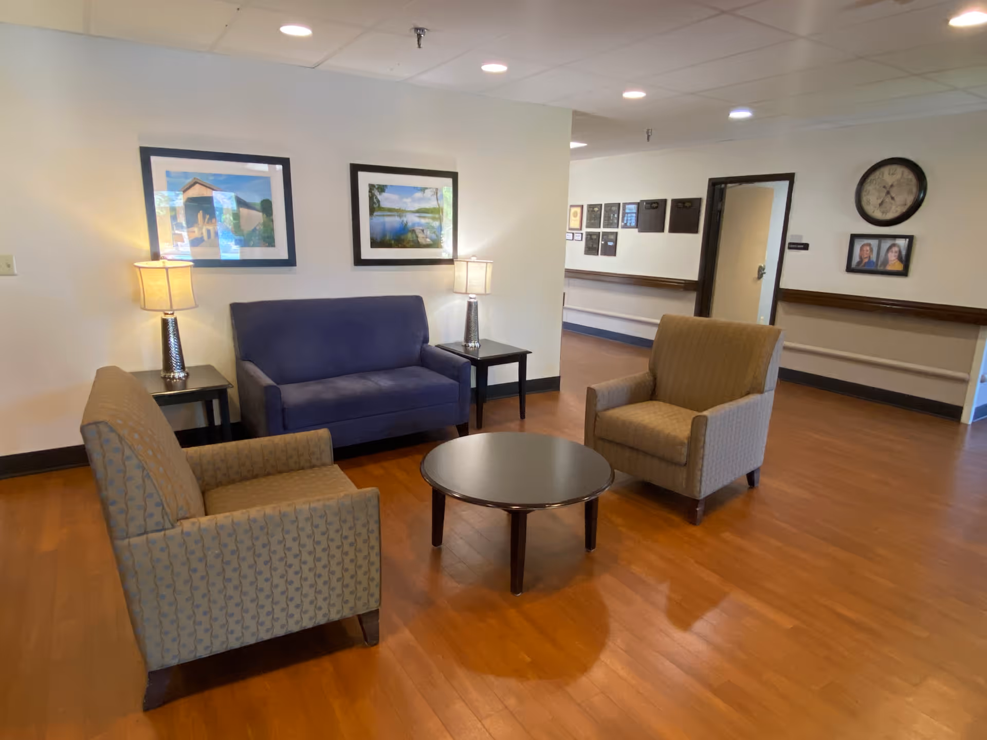 A seating area in a senior living facility with two beige armchairs and a blue loveseat arranged around a round wooden coffee table. Two table lamps with beige shades sit on small black side tables next to the loveseat. The walls are decorated with framed landscape photographs and a clock. The floor is wood, and there is a hallway visible in the background.