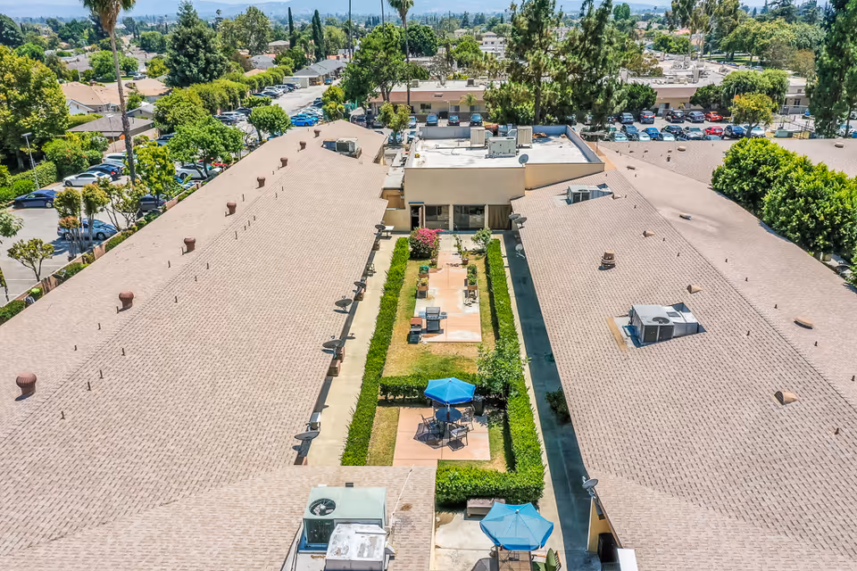 Aerial view of Santa Anita Assisted Living facility showing two long buildings with a central courtyard featuring green hedges, patio seating areas with blue umbrellas, and a walkway. Surrounding the buildings are trees, parked cars, and a residential neighborhood in the background.