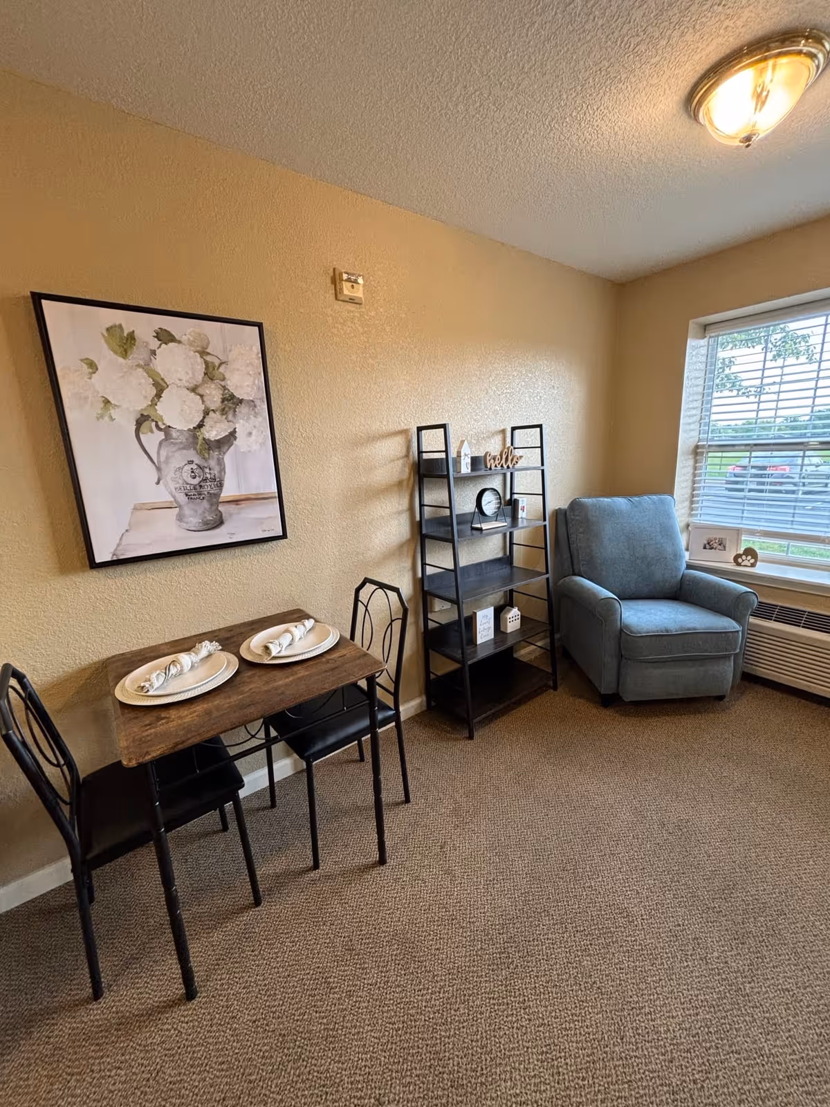 A small dining area with a wooden table set for two with plates and napkins, two black metal chairs, a black shelving unit with decorative items, a blue upholstered armchair, and a window with blinds showing a parking lot outside. A framed picture of white flowers in a vase hangs on the beige wall.