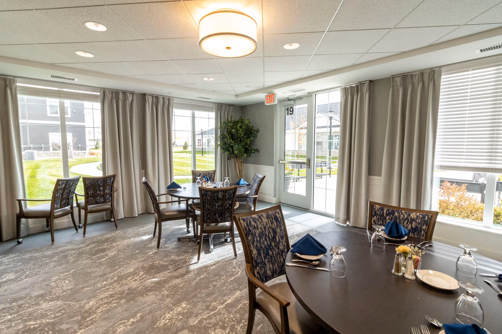 A bright dining room with round tables set with blue napkins, glasses, plates, and silverware. The room has large windows with beige curtains allowing natural light to fill the space. Outside, a green lawn and buildings are visible. There is a ceiling light fixture and an exit door with the number 19 above it.
