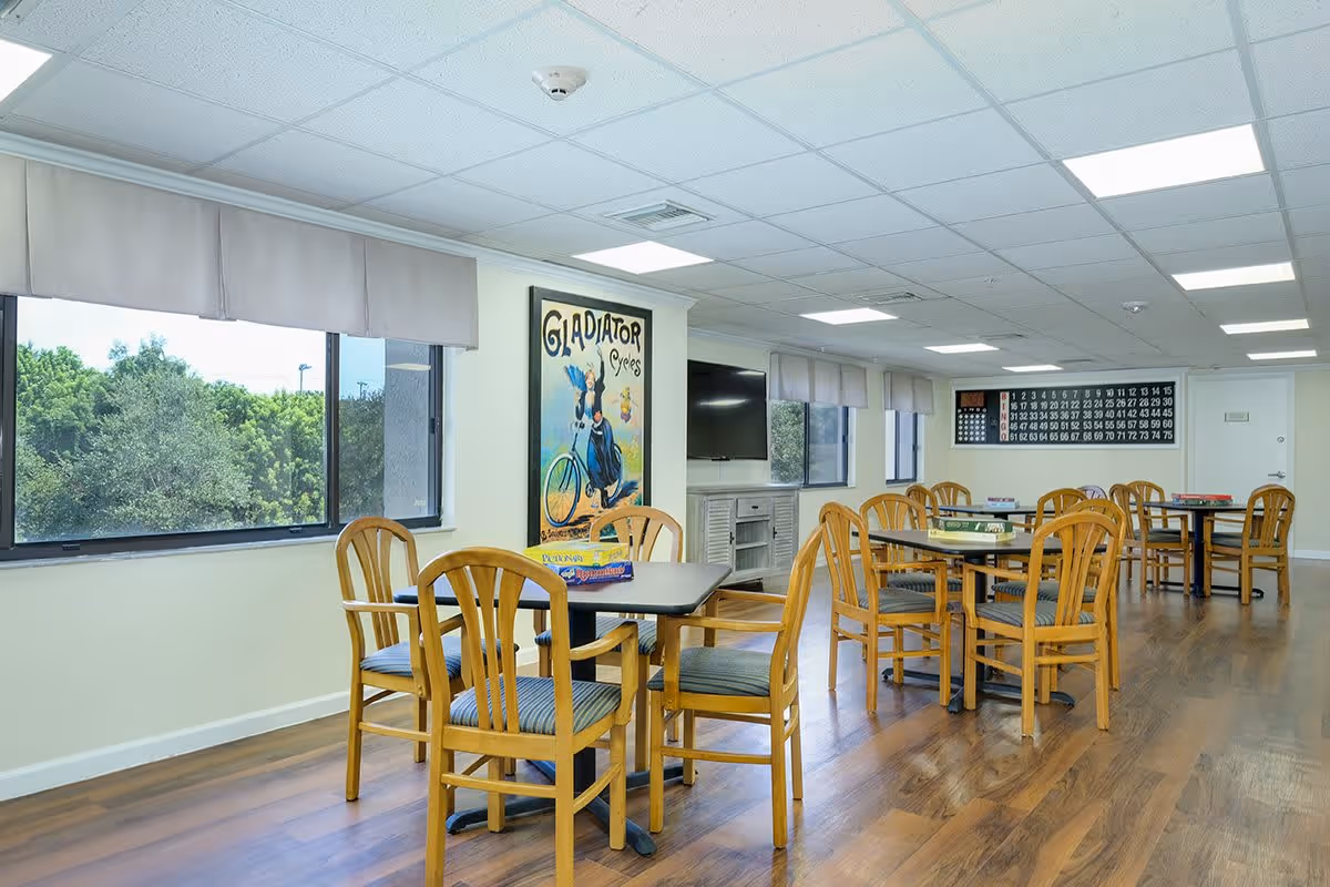 Bright communal dining/activity room with multiple tables and wooden chairs, large windows, a wall-mounted TV, and a bingo board on the wall.