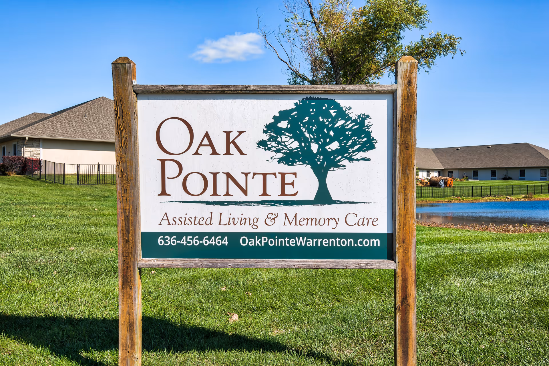 Wood-framed sign reading "Oak Pointe Assisted Living & Memory Care" on a grassy lawn with the facility buildings and a pond in the background.