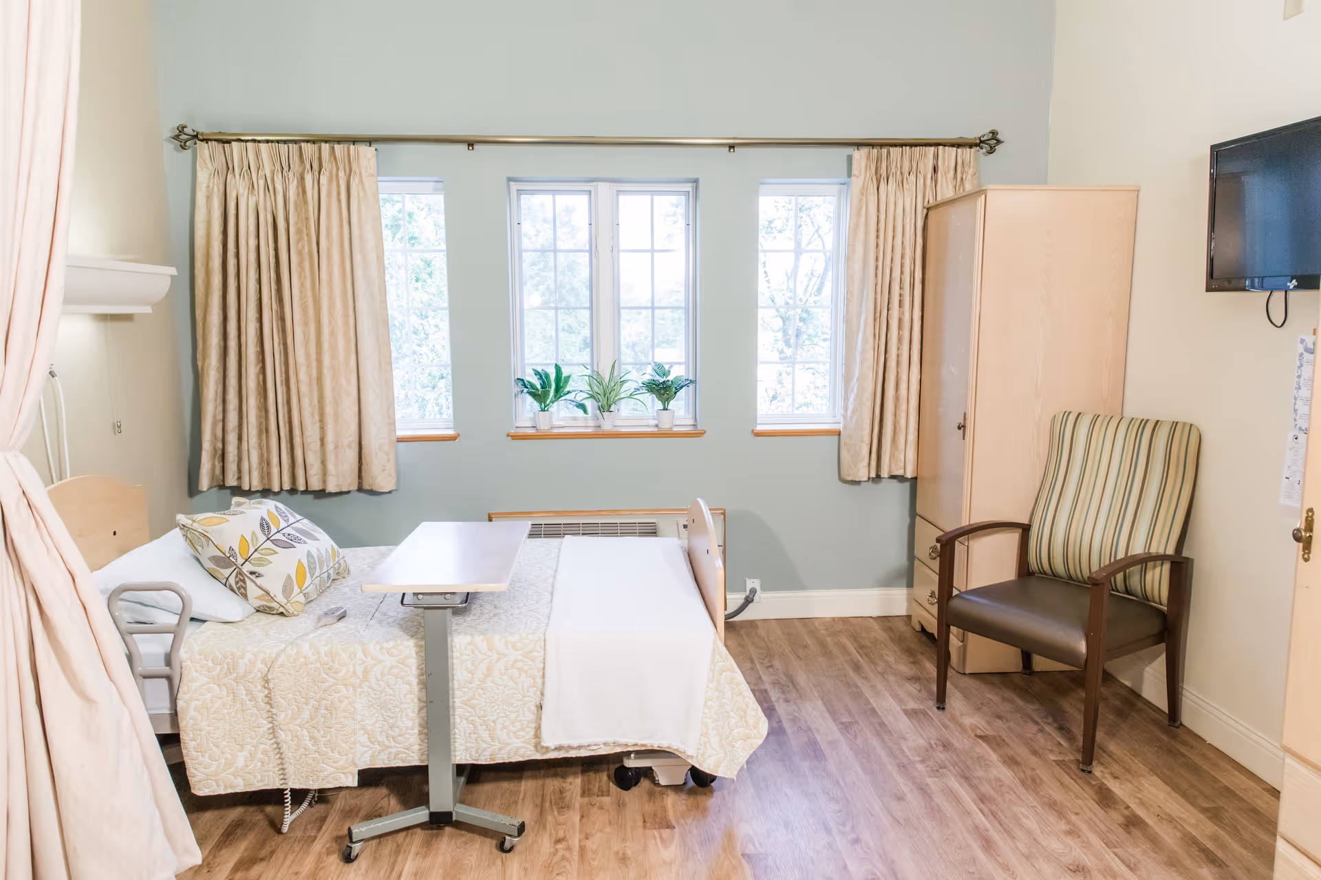A bright and clean nursing facility bedroom with a hospital bed covered in a beige patterned blanket and a decorative pillow. There is a small overbed table on wheels, a striped armchair, a wooden wardrobe, and a wall-mounted TV. Three windows with beige curtains let in natural light, and there are small potted plants on the windowsill. The room has wood flooring and light-colored walls.