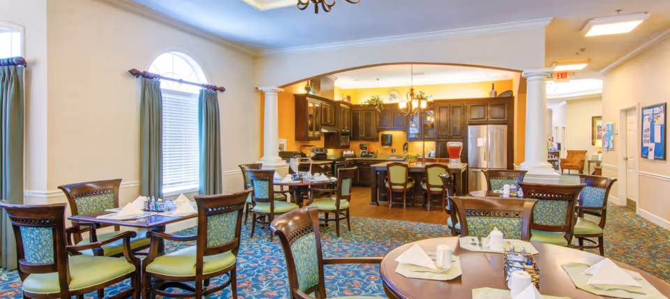 A bright and inviting dining area in a senior living facility featuring multiple wooden tables with green cushioned chairs. Each table is set with white napkins, cups, and condiments. The room has large windows with green curtains, a patterned blue carpet, and an open kitchen area with dark wooden cabinets and stainless steel appliances in the background. The space is well-lit with ceiling lights and a chandelier above the kitchen island.