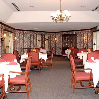 Formal dining room with tables set with white tablecloths, red upholstered chairs, a chandelier, and patterned wallpaper.