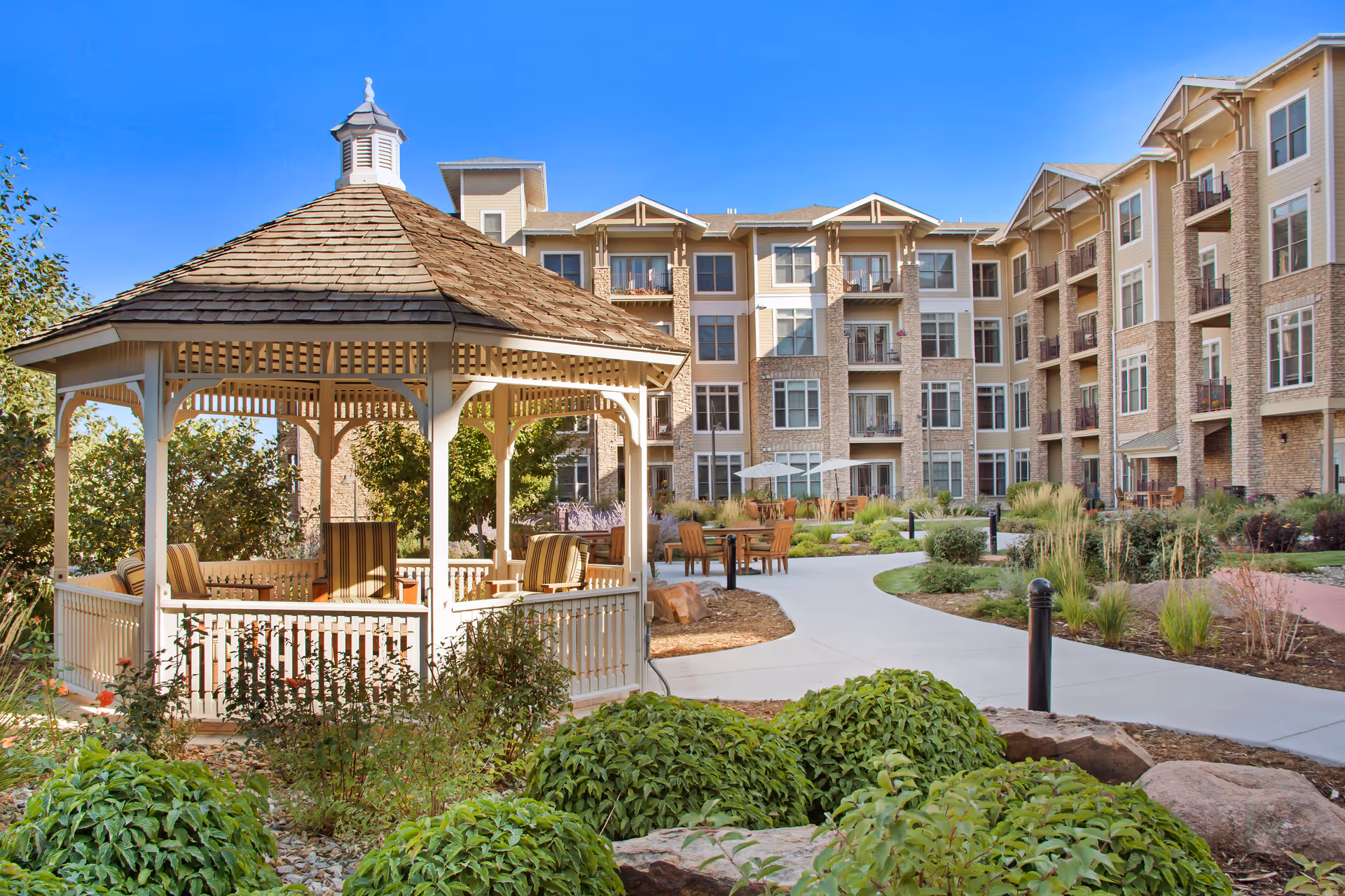 A wooden gazebo with seating in a landscaped courtyard in front of a multi-story senior living building under a blue sky.