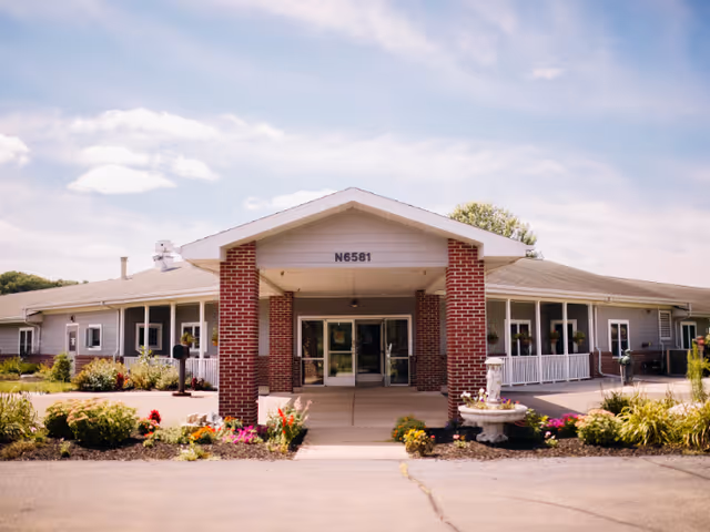 Front exterior view of a single-story senior living facility building with a covered entrance supported by brick columns, surrounded by landscaped flower beds and greenery under a partly cloudy sky.