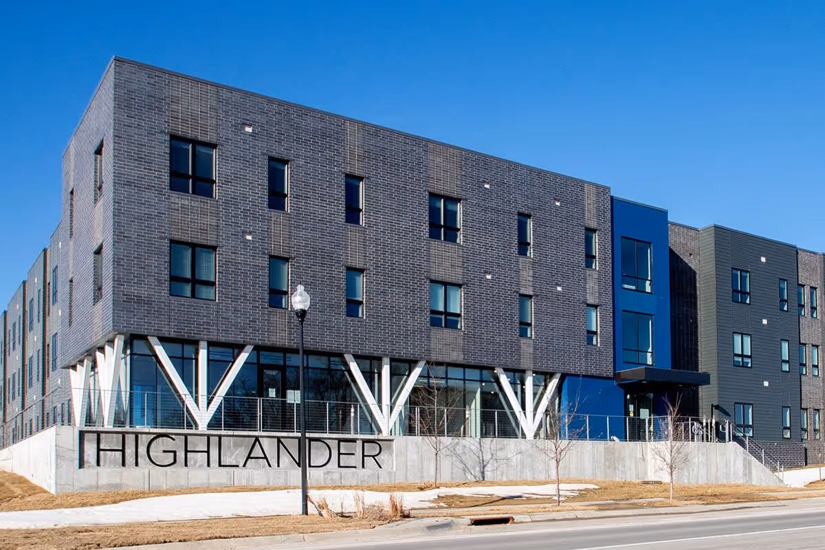 Exterior view of a modern three-story building with dark brick and blue paneling under a clear blue sky. The building has multiple windows and a concrete base with the word 'HIGHLANDER' displayed on it. There is a street and sidewalk in the foreground with a streetlamp and some small leafless trees.