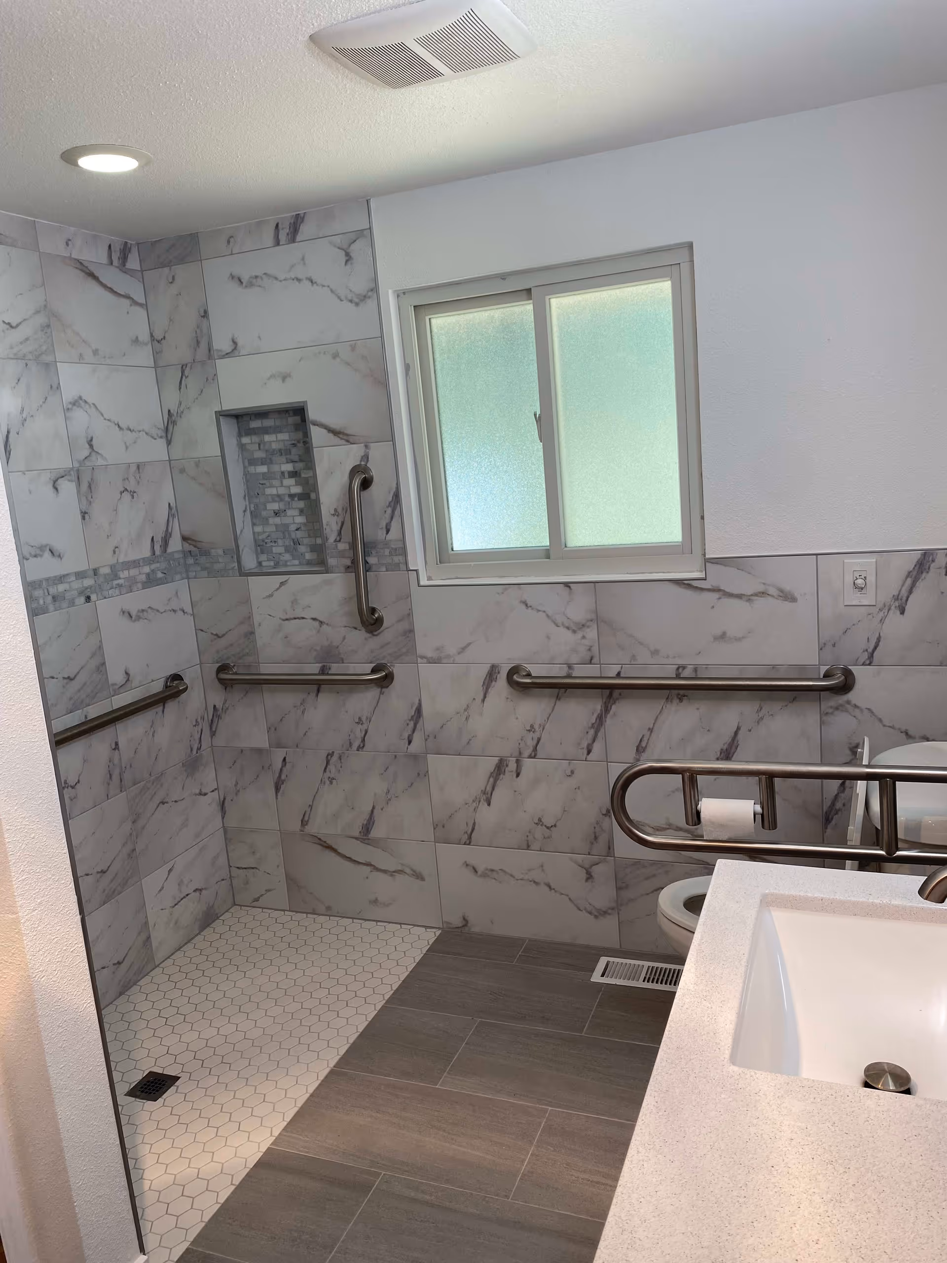 A modern bathroom with a walk-in shower featuring marble-patterned tiles and multiple stainless steel grab bars. The shower floor has small hexagonal white tiles, and there is a recessed shelf in the shower wall. A frosted window is above the toilet, which also has a grab bar with a toilet paper holder. A white sink countertop is visible in the foreground.