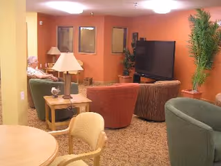 A cozy living room area with several upholstered chairs arranged around a large flat-screen TV. There is a wooden side table with a lamp and a small decorative item. A person is seated on one of the chairs near the back wall, which is painted orange. There are also some green plants in the room, and the carpet has a patterned design.