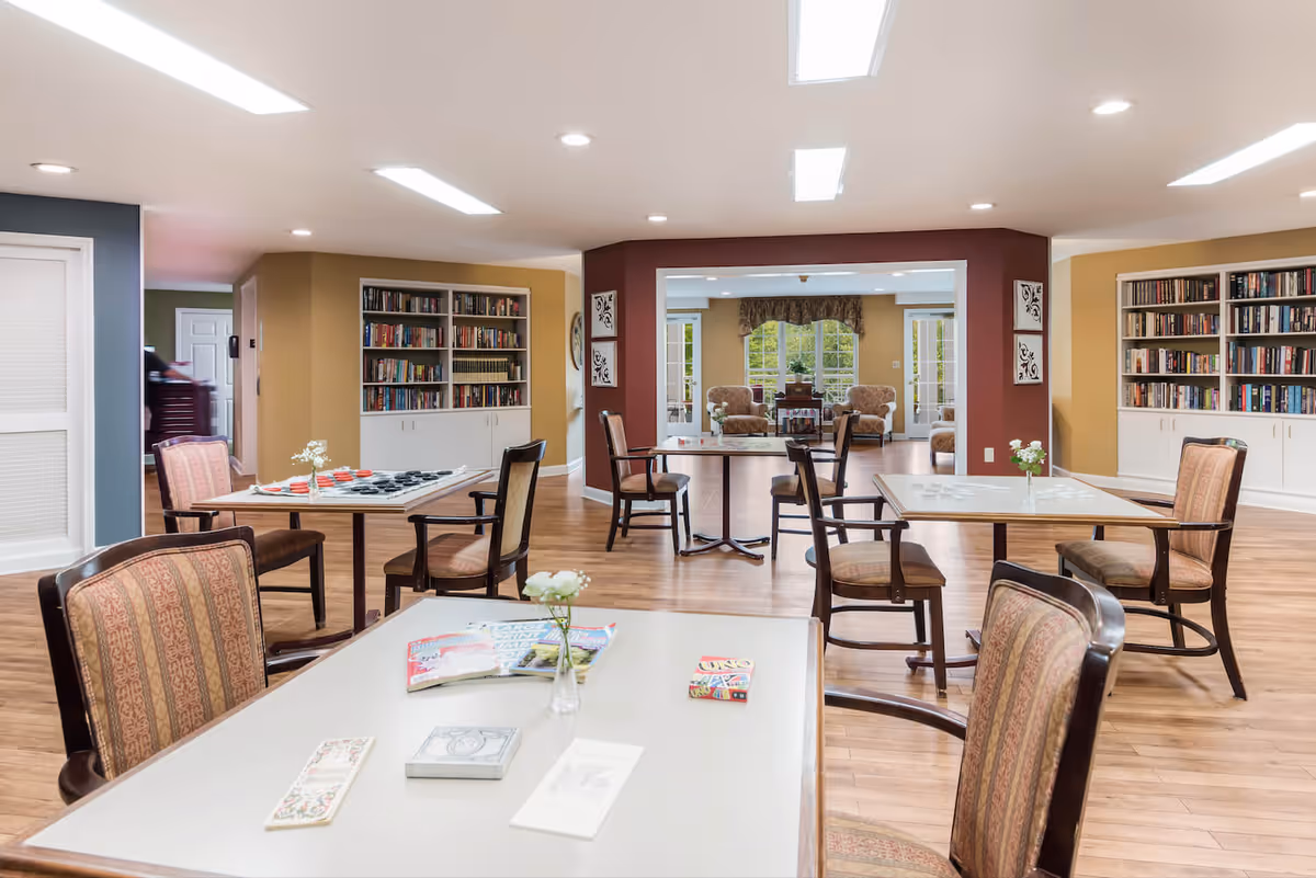 Bright communal activity room with tables and chairs, bookshelves along the walls, and a seating area by windows.
