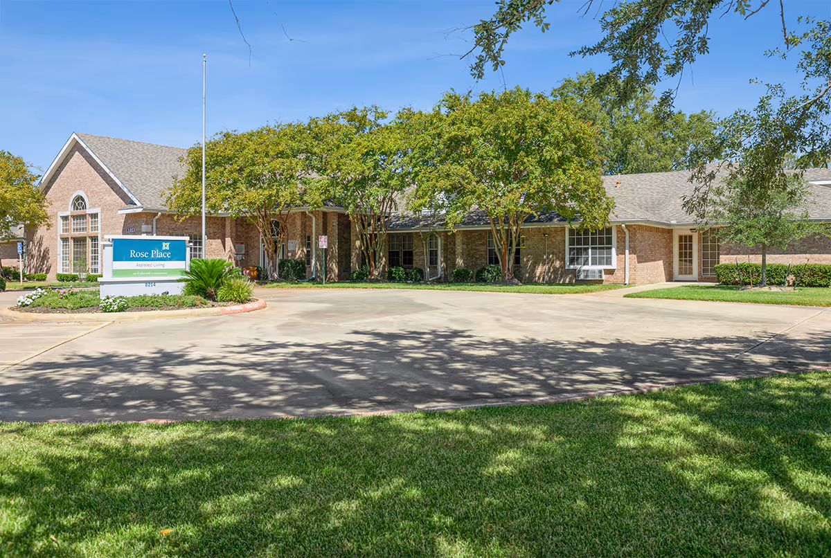 Exterior view of a single-story brick building with a pitched roof, surrounded by trees and greenery under a clear blue sky. A sign in front reads 'Rose Place Assisted Living'. There is a paved driveway and well-maintained lawn in the foreground.