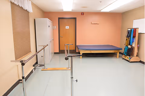A therapy or exercise room with parallel bars for walking support, a padded therapy table against the far wall, a white cabinet, a door, and a rack holding various exercise bands and equipment. The walls are painted beige and peach, and the floor is light-colored linoleum.