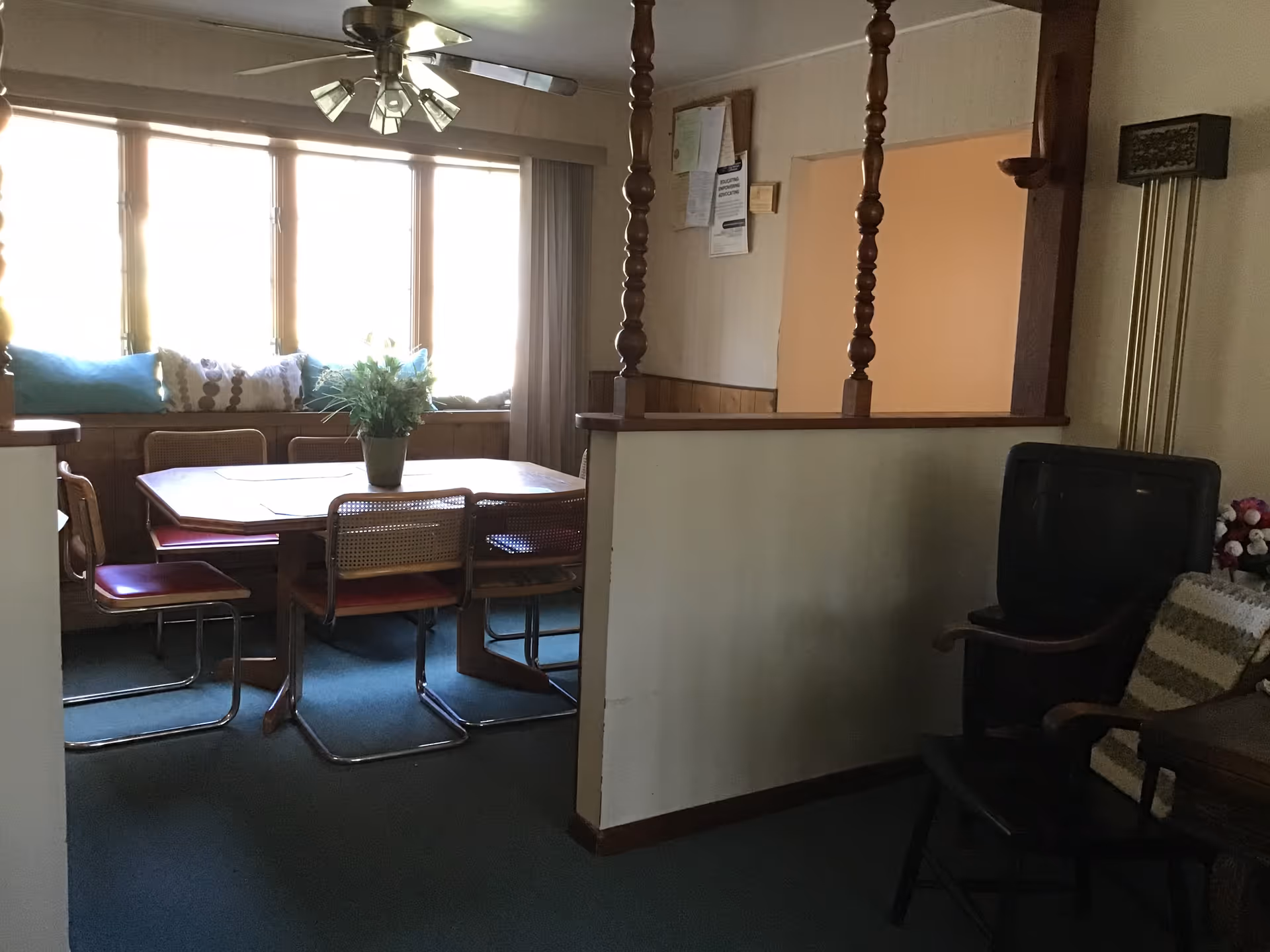 Interior view of a dining area with a wooden table surrounded by six chairs, a ceiling fan with lights above, and a window seat with cushions in front of large windows letting in bright light. To the right, there is a small sitting area with a black chair, a wooden chair with a cushion, and a tall floor lamp.