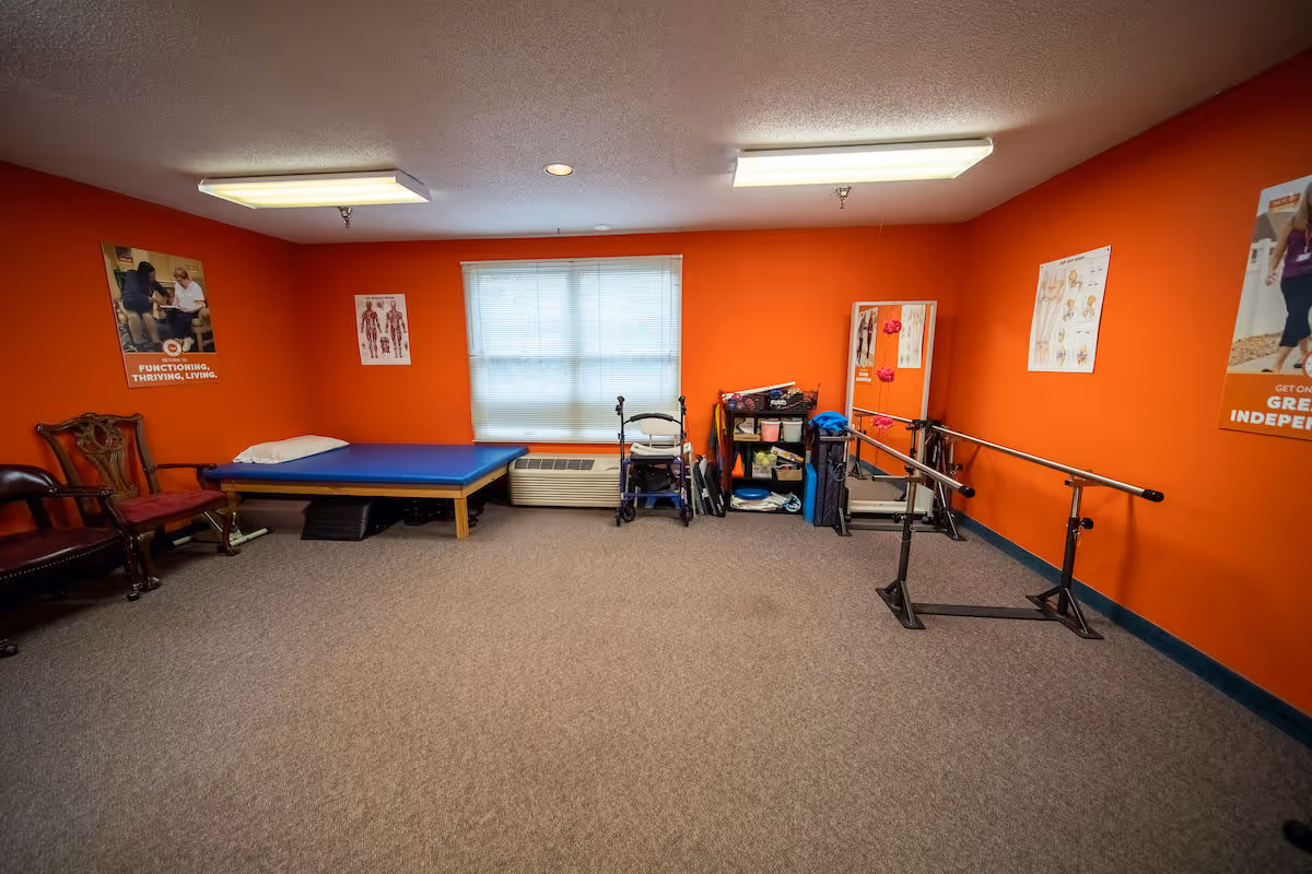 A therapy room with bright orange walls, featuring a blue padded therapy table with a pillow, two wooden chairs with red cushions, a walker, a shelf with various therapy supplies, a large mirror, and parallel bars for walking exercises. Anatomical posters and motivational posters are displayed on the walls.