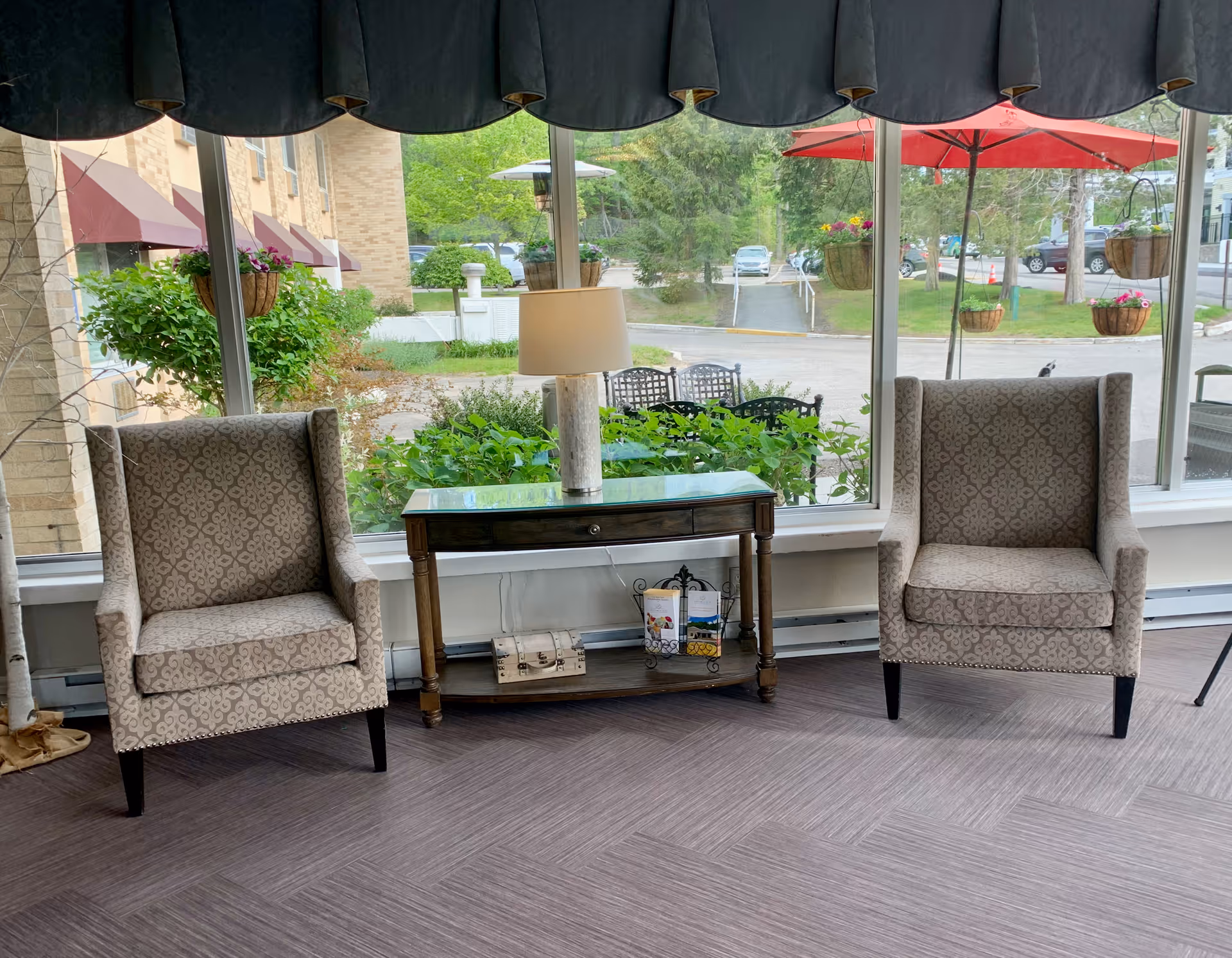 Two patterned armchairs flank a wooden console table with a lamp in a seating area by large windows overlooking a courtyard with umbrellas and hanging planters.
