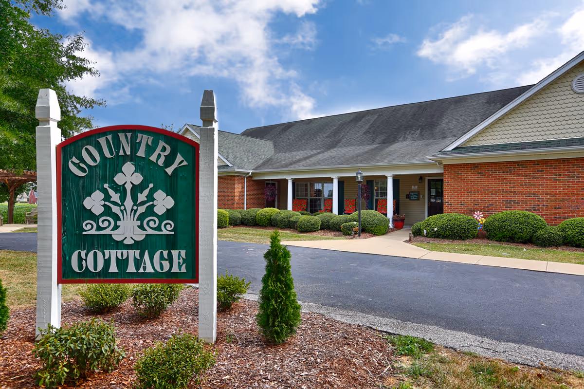 Exterior view of Country Cottage Lawrenceburg facility showing a green and white wooden sign with the facility name in front of a brick building with a gray roof, surrounded by bushes and a paved driveway under a partly cloudy sky.