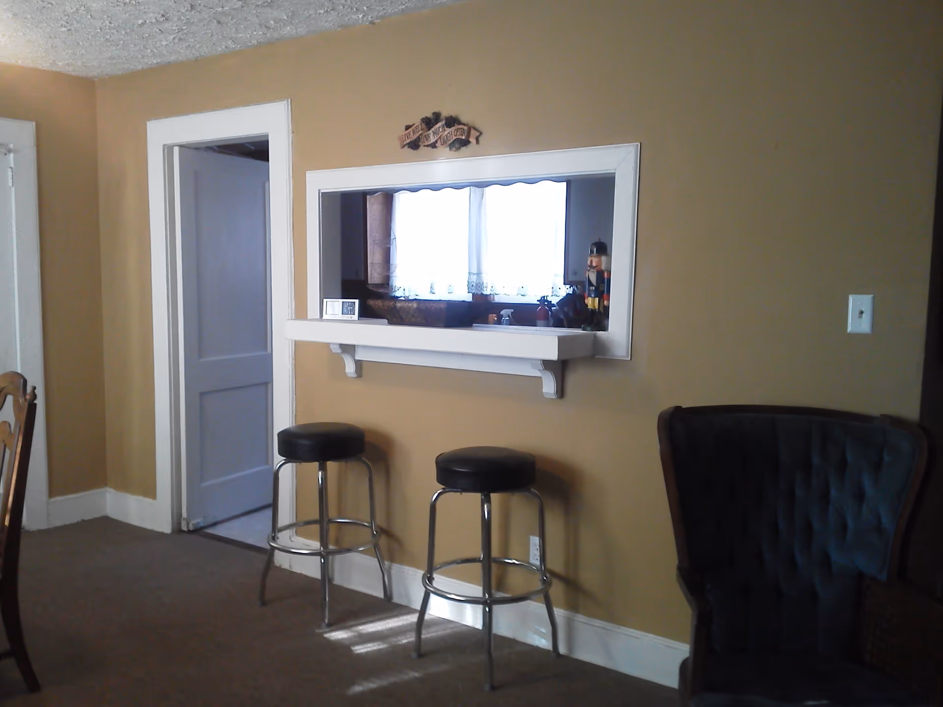 Room with a countertop pass-through to a kitchen, two metal bar stools, and an upholstered armchair.