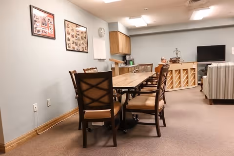 Communal activity room with a long wooden table and chairs, a piano and TV, a small kitchenette and framed photos on the wall.