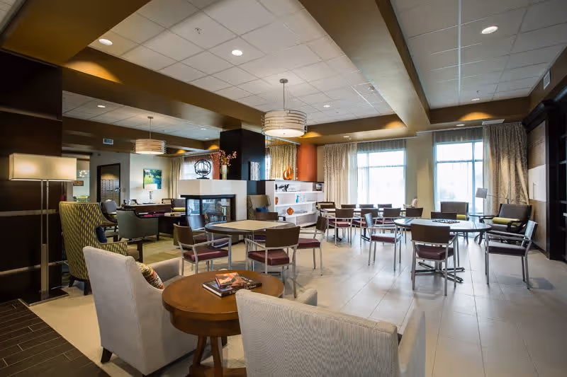 A spacious and well-lit common area with multiple tables and chairs arranged for seating. The room features large windows with curtains allowing natural light, modern ceiling lights, and a central fireplace with decorative items on shelves. Comfortable armchairs and a small round wooden table with books are also visible.