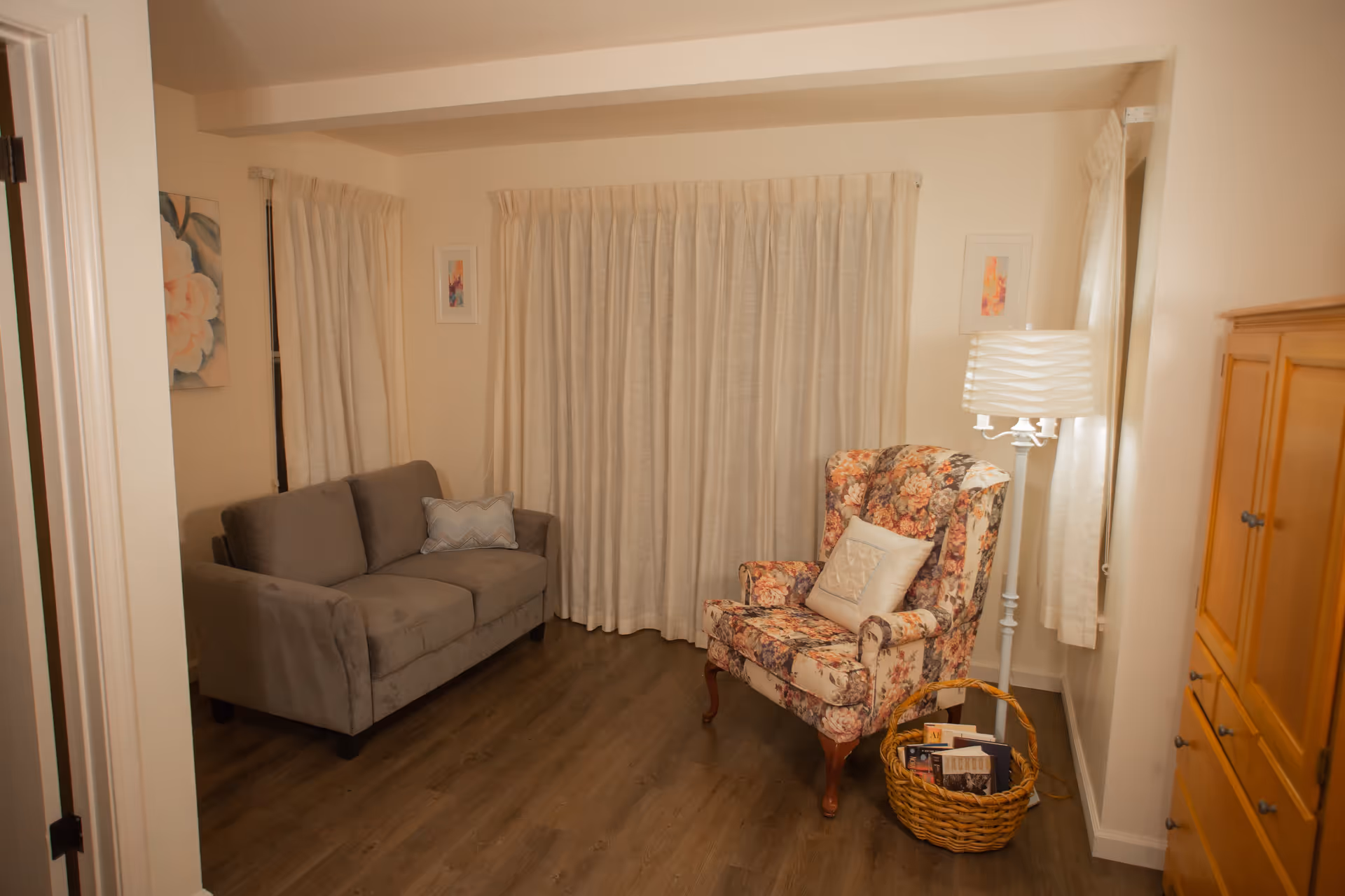 A cozy living room with a gray sofa on the left and a floral upholstered armchair with a white pillow on the right. A white floor lamp stands behind the armchair, next to a basket filled with books. The room has light-colored curtains covering the windows and light wood flooring. There is a wooden cabinet on the right side of the image and framed artwork on the walls.