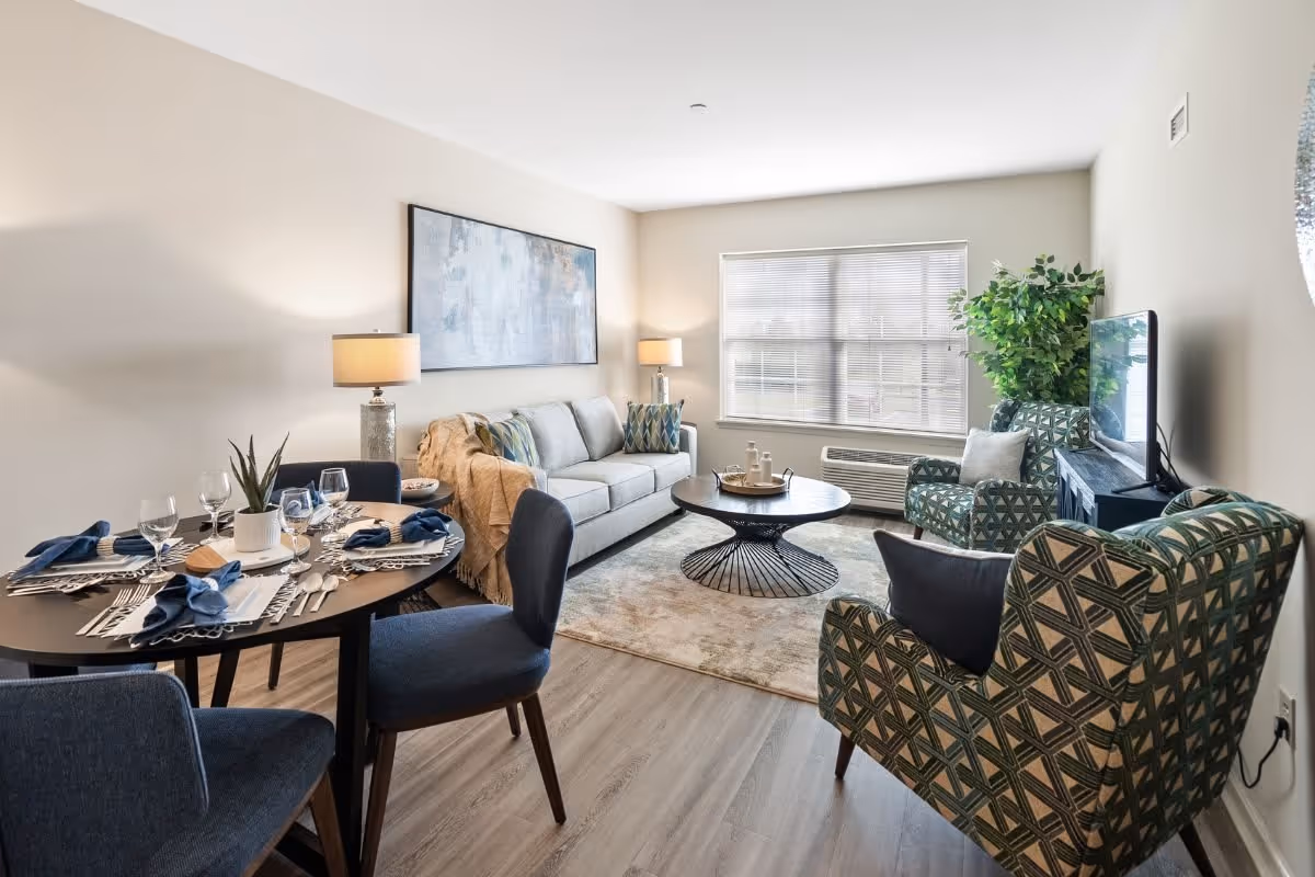 A bright and modern living room and dining area featuring a round dining table set with plates, glasses, and blue napkins, a light gray sofa with decorative pillows and a throw blanket, two patterned armchairs, a round coffee table on a beige rug, a large window with blinds, two table lamps, a large abstract painting on the wall, and a flat-screen TV on a stand with a tall green plant beside it.