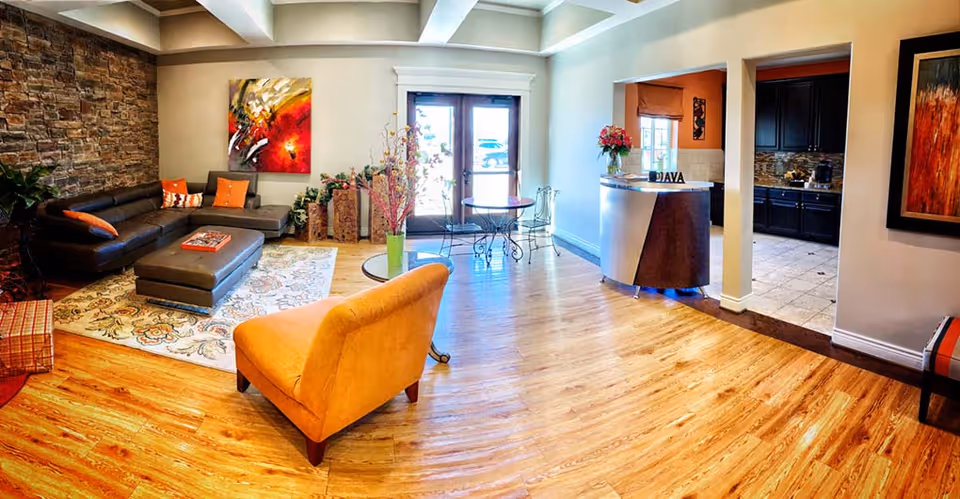 Bright assisted-living lounge with hardwood floors, a sectional sofa, an orange accent chair, artwork, and a small reception desk.