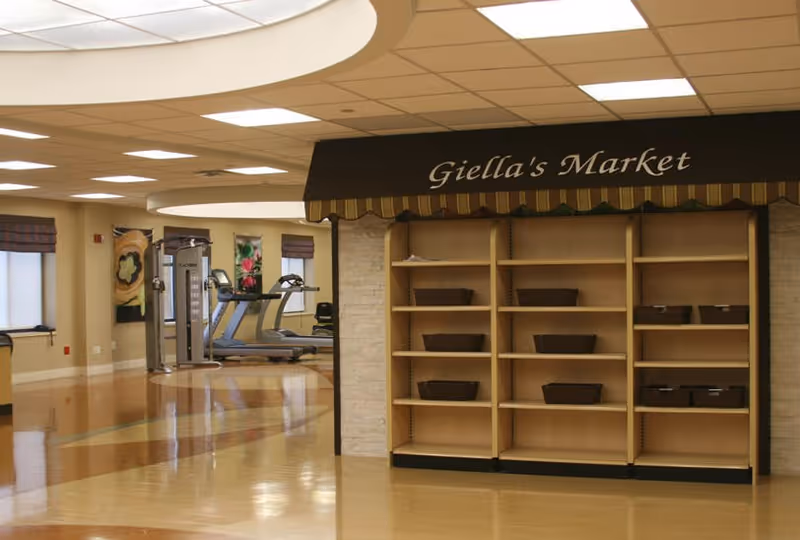 Interior view of a senior living facility showing an empty market area labeled 'Giella's Market' with shelves and baskets, and in the background, exercise equipment including treadmills and weight machines.