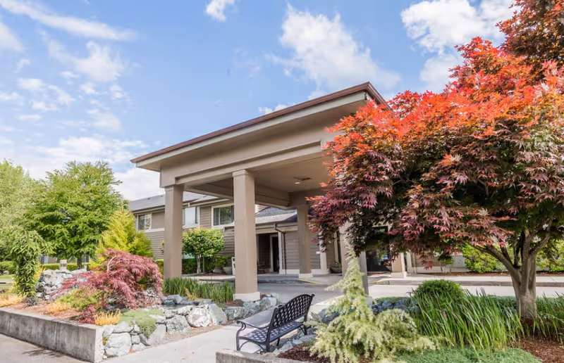 Senior living facility exterior with a covered entrance, bench, and landscaped trees.