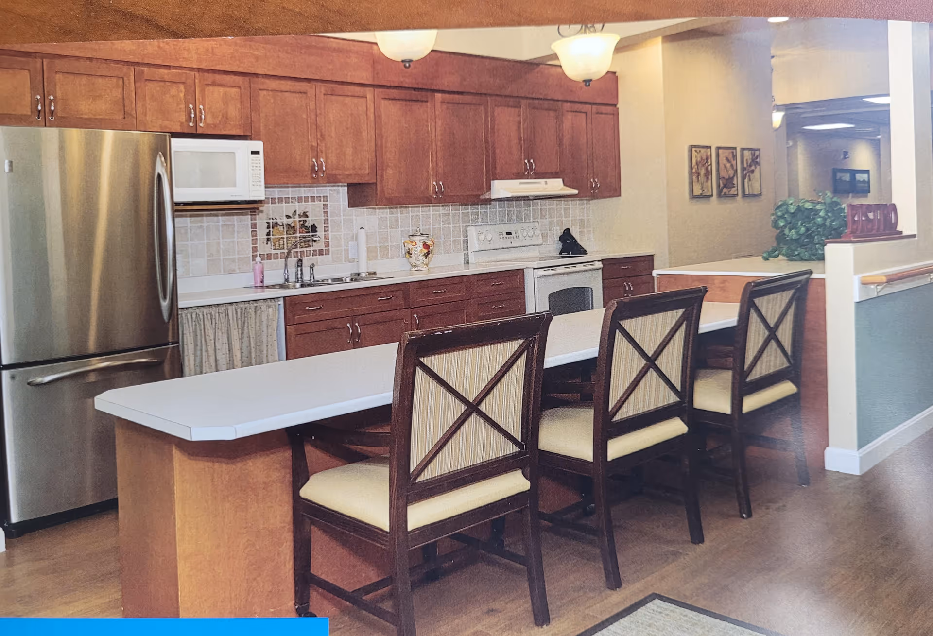 A kitchen area in a senior living facility featuring wooden cabinets, a stainless steel refrigerator, a microwave, an electric stove, and a long white countertop with four wooden chairs with cushioned seats. The kitchen has tiled backsplash with a decorative tile centerpiece and overhead lighting fixtures. In the background, there is a hallway with framed artwork and a green plant on a counter.