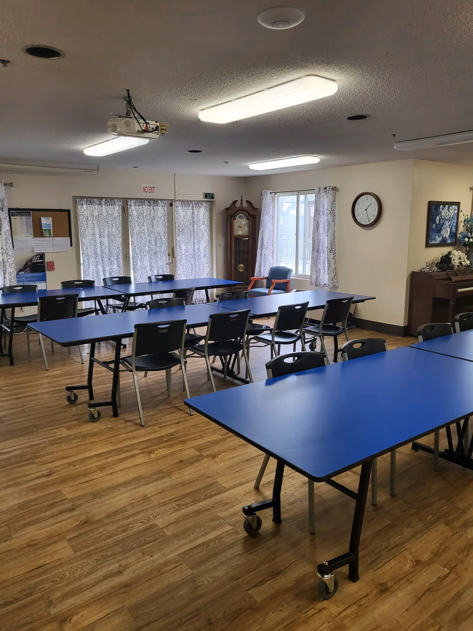 A well-lit room with multiple blue tables and black chairs arranged in rows on a wooden floor. The room has white walls with patterned curtains on the windows and a clock on the wall. There is a grandfather clock in the corner, a bulletin board, and a piano with floral decorations. Ceiling lights and a projector are visible overhead.