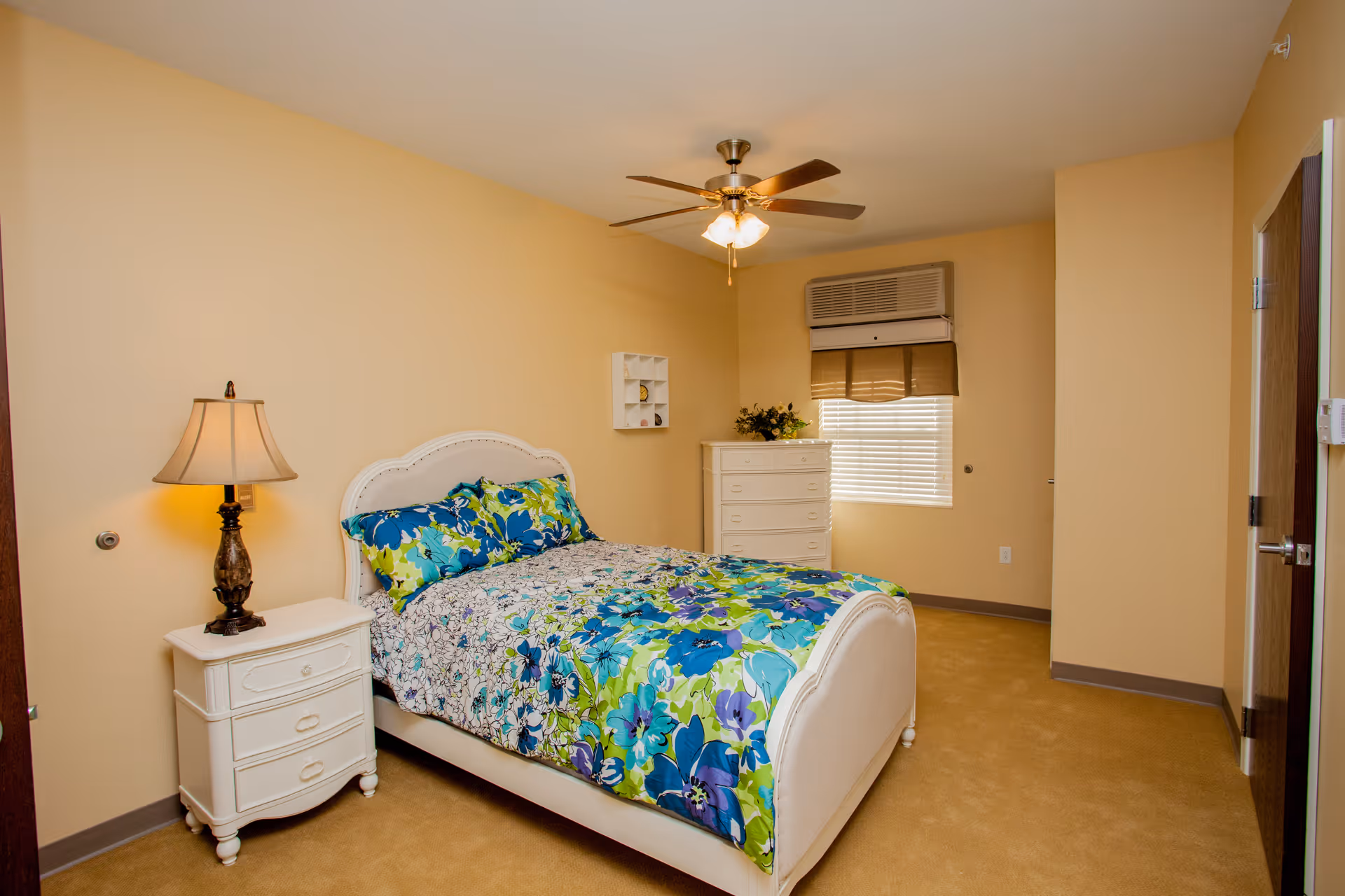 A bedroom with beige walls and carpet featuring a white bed with a colorful floral bedspread in shades of blue, green, and purple. Next to the bed is a white nightstand with a decorative lamp. A white dresser with flowers on top is positioned under a window with blinds and a brown valance. A ceiling fan with lights is mounted on the ceiling.
