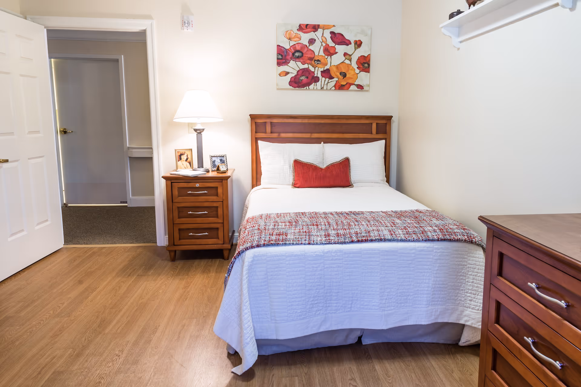 A neatly made bed with white bedding and a red decorative pillow in a senior living facility bedroom. There is a wooden nightstand with a lamp, framed photos, and a book next to the bed. A colorful floral painting hangs on the wall above the headboard. The room has wooden flooring and light-colored walls, with an open door leading to a hallway.