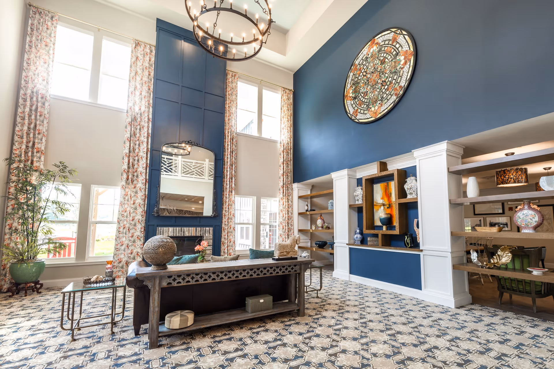 A spacious and elegant living room with high ceilings and large windows covered by floral curtains. The room features a dark blue accent wall with a large decorative circular piece and built-in shelves displaying various vases and art. A fireplace with a mirror above it is centered on another wall. The floor is covered with a patterned carpet, and there is a wooden console table behind a leather sofa with decorative items on it. A large chandelier hangs from the ceiling.
