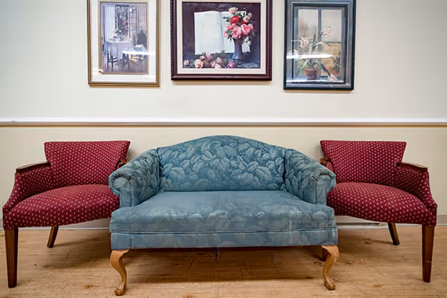 Small seating area with a blue upholstered loveseat flanked by two red armchairs and three framed paintings on the wall.