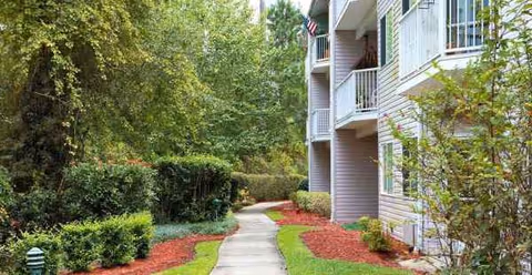 A curved paved walkway beside a multi-story senior living building with trimmed shrubs.