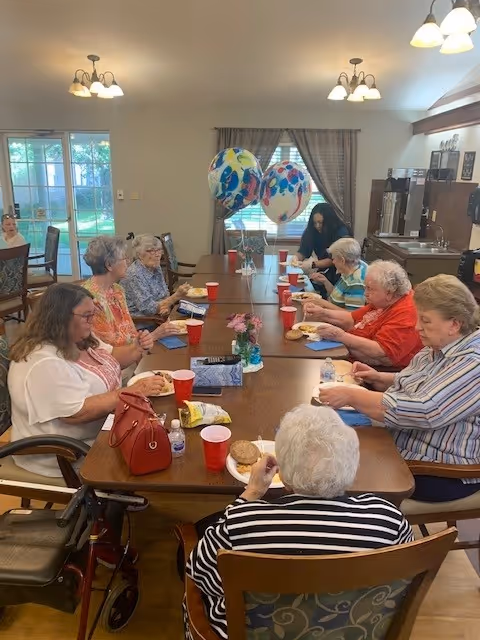 Several elderly residents seated around a long dining table eating and talking in a communal dining room with balloons.