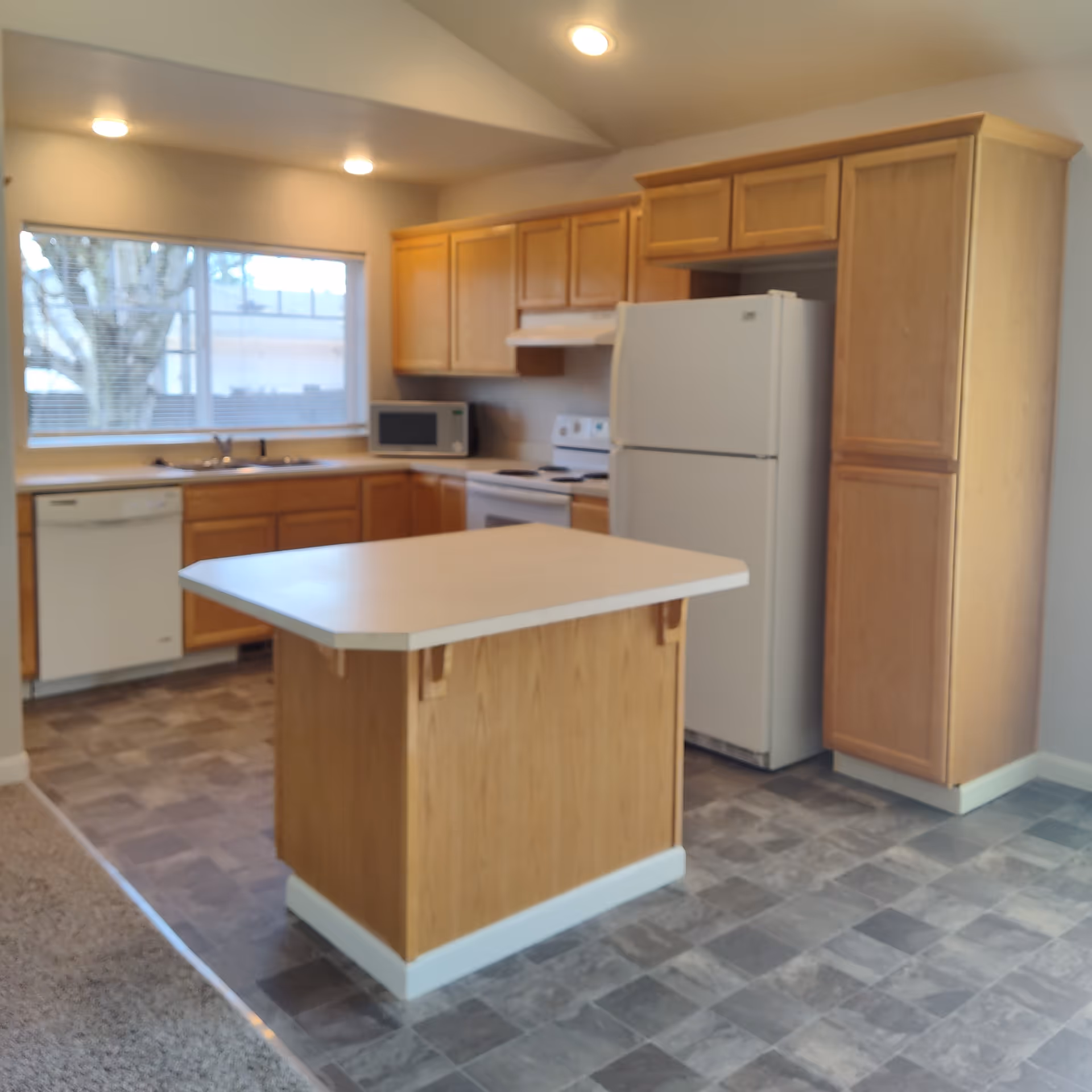 Bright kitchen with a central island, wooden cabinets, white refrigerator, stove, dishwasher, and a window above the sink.