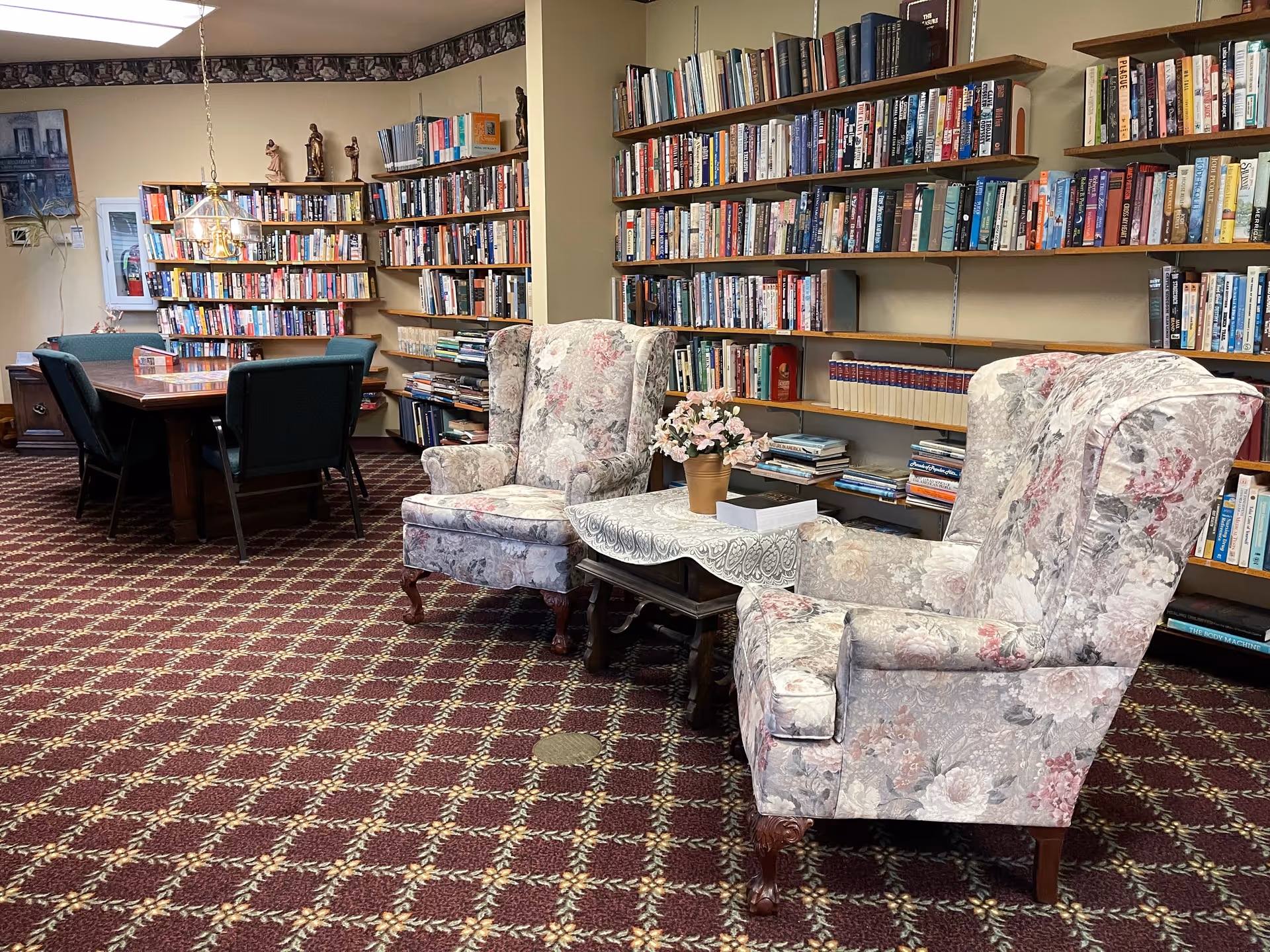 A cozy library room with floral upholstered armchairs and a small table with a flower pot in the foreground. The walls are lined with shelves filled with books. In the background, there is a wooden table with green chairs around it and a hanging light fixture above the table. The floor is covered with a patterned carpet.