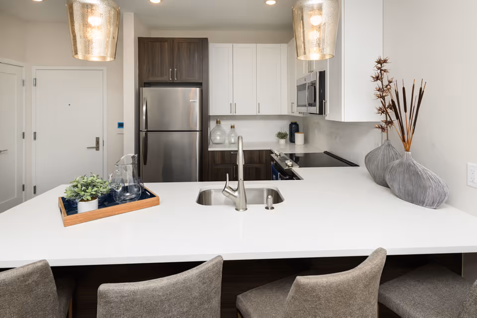 Modern kitchen with a white countertop island featuring a built-in sink and faucet. Three gray upholstered chairs are positioned at the island. The kitchen has stainless steel appliances including a refrigerator and microwave, white and dark wood cabinetry, two pendant lights hanging above the island, and decorative vases with dried plants on the counter.