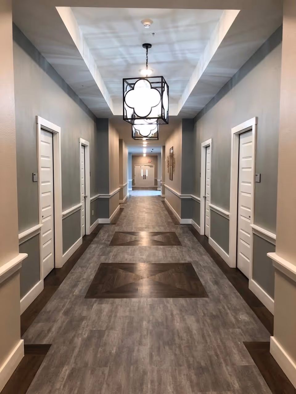 A long, well-lit hallway with gray walls and white doors on both sides. The floor has a patterned design with darker wood inlays. Modern square pendant lights with a clover-shaped design hang from the ceiling. At the end of the hallway, double doors are visible.