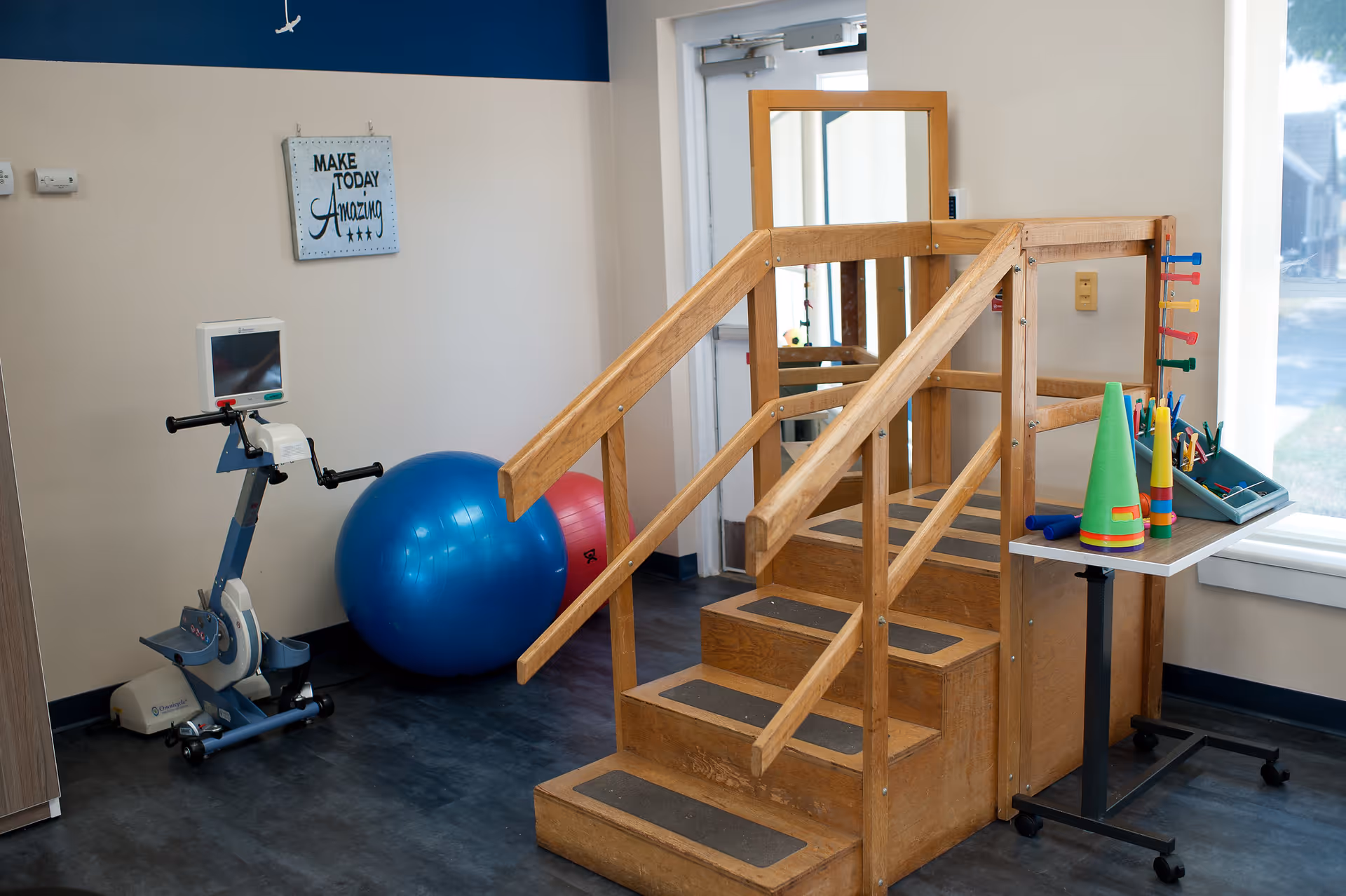 Physical therapy room with wooden practice stairs, an exercise bike, large stability balls and therapy tools by a window.