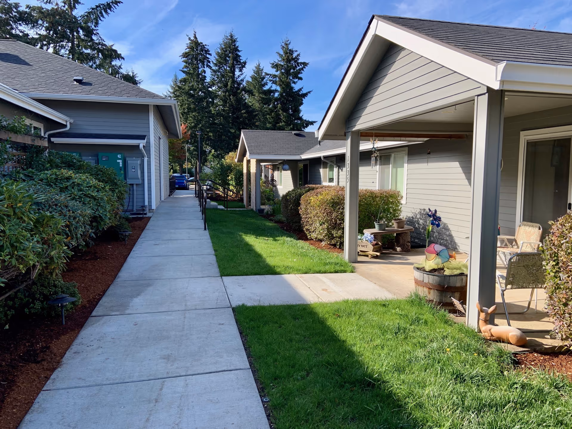 Concrete sidewalk between single-story senior living cottages with covered porches, lawn, shrubs, and garden decorations under a blue sky.