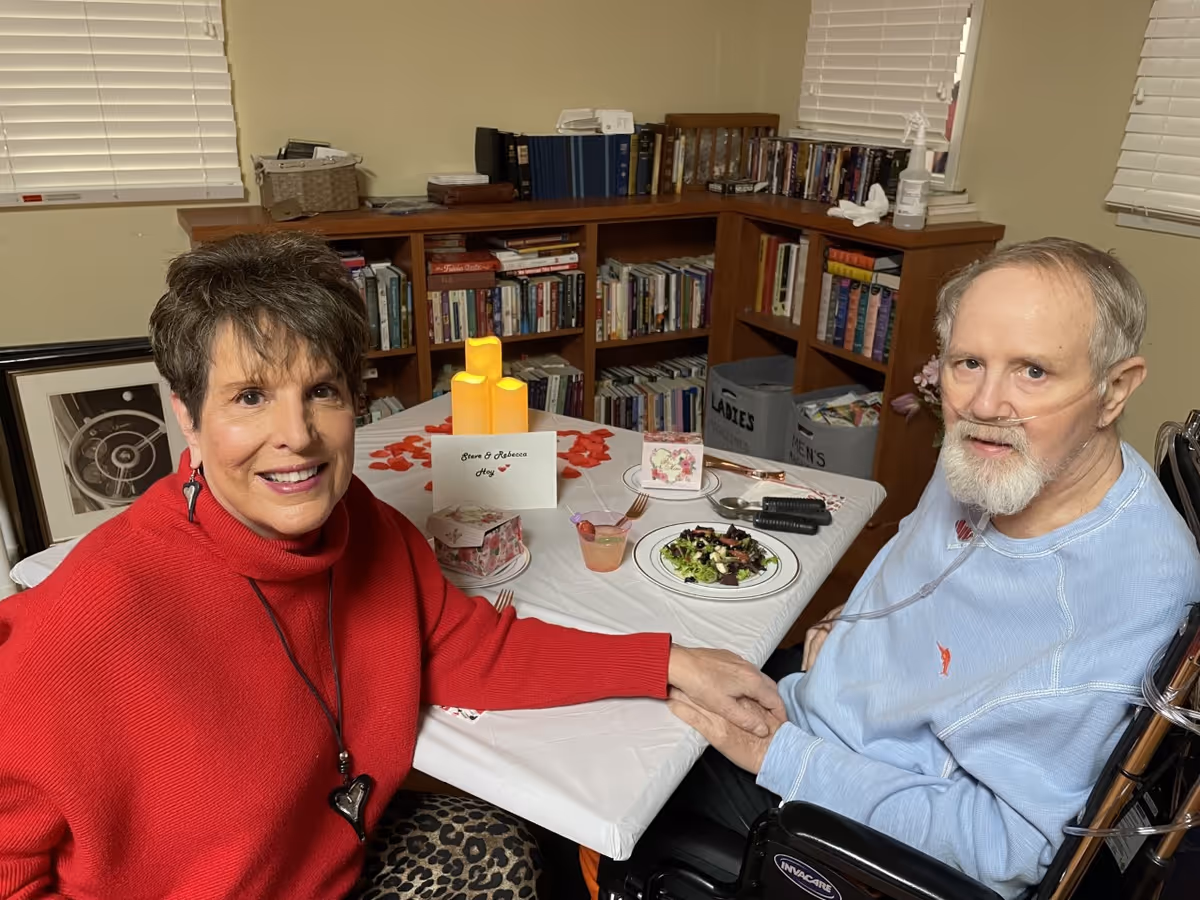 An elderly man in a wheelchair with an oxygen tube and a woman in a red sweater holding hands across a table set with a salad, candles, and a card. They are in a room with bookshelves filled with books and windows with blinds.
